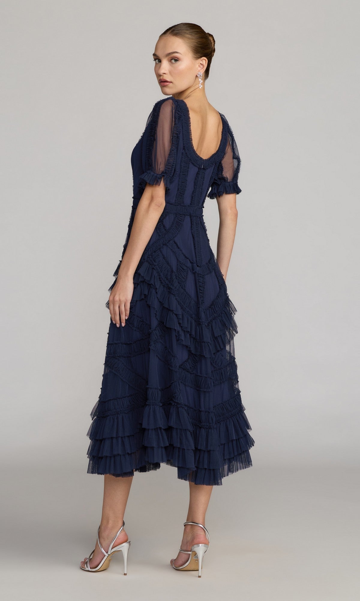 A woman stands sideways in the Mac Duggal 8248 Midi Wedding Guest Dress—a navy blue, short-sleeve, ruffled midi dress—paired with silver high-heeled sandals, glancing over her shoulder against a plain background.